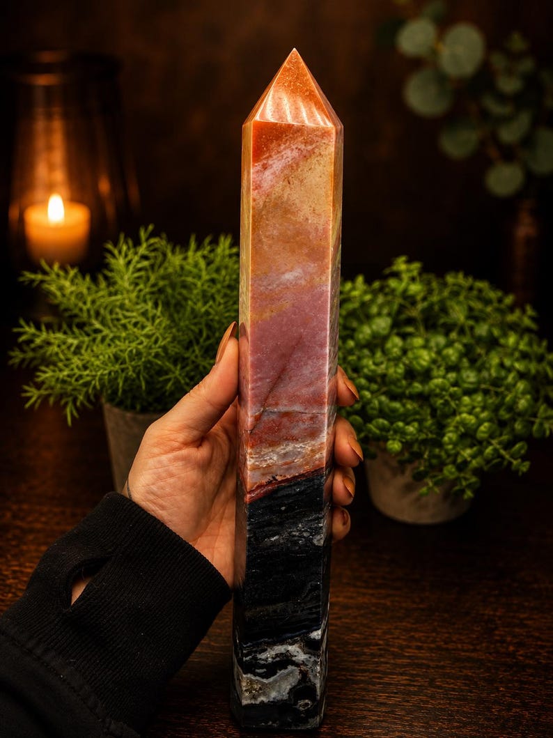 May include: A tall, polished crystal tower featuring a multi-colored gradient. The stone transitions from a deep black base through layers of dark red and earthy brown, ending in a lighter, translucent orange-toned point at the top.