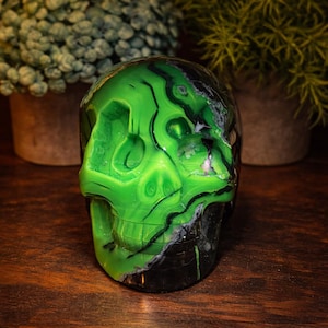 May include: A decorative skull sculpture with a vibrant green and black color scheme. The skull is made of a polished material, with intricate details and a glossy finish. The sculpture is displayed on a wooden surface.