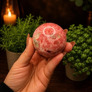 May include: A polished spherical stone featuring a mottled pattern of deep pink, red, and muted green mineral inclusions. The surface of the sphere is smooth and reflective, displaying natural colour variations throughout its rounded, solid form.