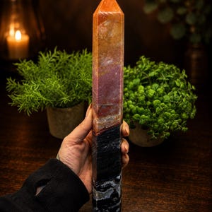 May include: A tall, polished crystal tower with a hexagonal shape. The stone features a gradient of colors, transitioning from a warm orange and reddish-brown at the pointed top to a deep, solid black with white mineral inclusions at the base.