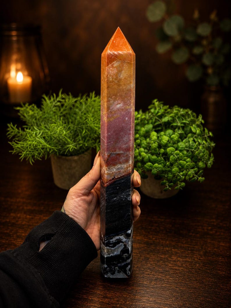 May include: A tall, polished crystal tower with a hexagonal shape. The stone features a gradient of colors, transitioning from a warm orange and reddish-brown at the pointed top to a deep, solid black with white mineral inclusions at the base.