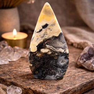 May include: A teardrop-shaped stone with a polished surface. The stone features a blend of colors, including yellow, black, gray, and cream, with intricate patterns. The stone is displayed on a brown surface, suggesting a decorative or collectible item.