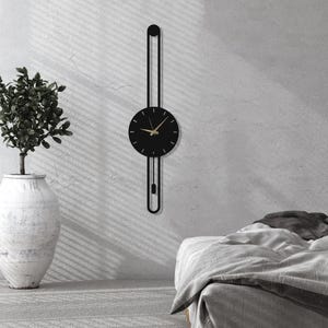 May include: A modern black wall clock with gold hands and a pendulum design. The clock is mounted on a light gray wall, with a plant in a white vase to the left and a bed on the right. The clock's design is minimalist and elegant.