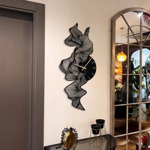 May include: Black wall clock with a distinctive design. The clock has a circular face with Roman numerals and gold hands, integrated into a flowing, abstract shape. The clock is mounted on a white wall.