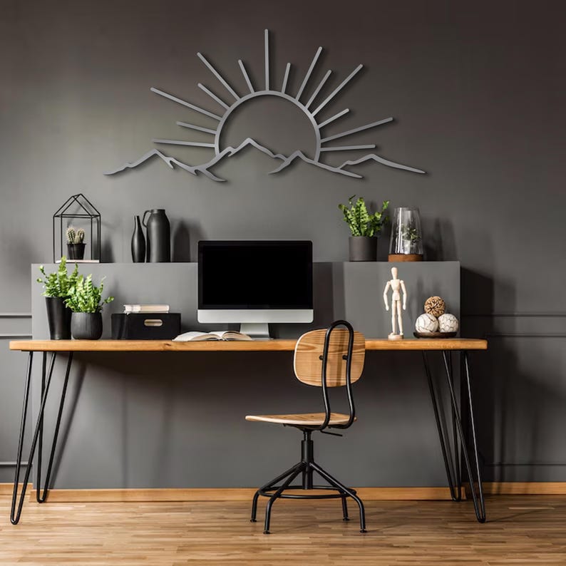 Pu&ograve; includere: Un moderno allestimento per l'home office con una scrivania in legno, gambe a forcina nere e una sedia nera. Un'opera d'arte da parete in metallo con sole e montagne &egrave; appesa sopra un monitor del computer. Piante e accessori decorativi completano lo spazio di lavoro.