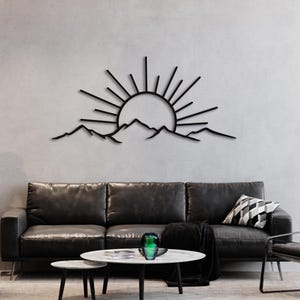 Pu&ograve; includere: Arte da parete in metallo nero raffigurante un'alba sulle montagne. I raggi del sole si estendono da un cerchio centrale, con una catena montuosa sottostante. L'opera d'arte &egrave; montata su una parete grigio chiaro sopra un divano in pelle nera.