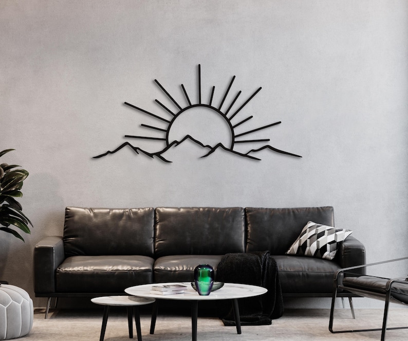 Pu&ograve; includere: Arte da parete in metallo nero raffigurante un'alba sulle montagne. I raggi del sole si estendono da un cerchio centrale, con una catena montuosa sottostante. L'opera d'arte &egrave; montata su una parete grigio chiaro sopra un divano in pelle nera.