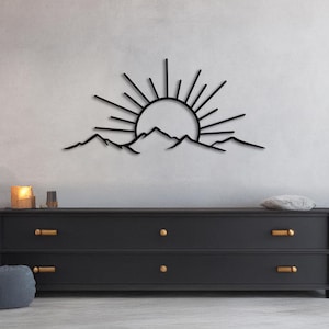 Pu&ograve; includere: Opera d'arte da parete in metallo nero raffigurante un'alba su una catena montuosa. Il sole ha linee radianti e le montagne hanno una silhouette frastagliata. L'opera &egrave; montata su una parete grigio chiaro sopra un com&ograve; nero con ferramenta dorata.