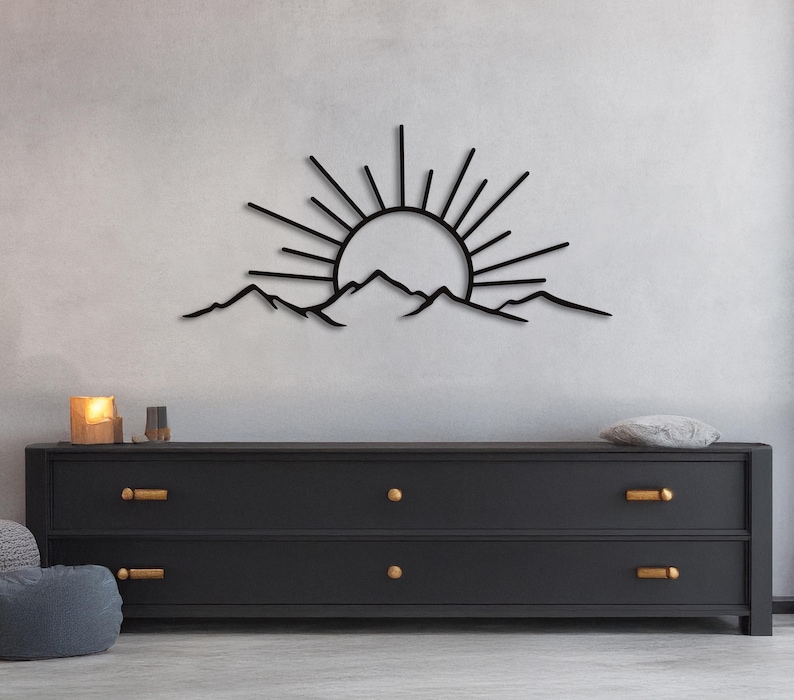 Pu&ograve; includere: Opera d'arte da parete in metallo nero raffigurante un'alba su una catena montuosa. Il sole ha linee radianti e le montagne hanno una silhouette frastagliata. L'opera &egrave; montata su una parete grigio chiaro sopra un com&ograve; nero con ferramenta dorata.