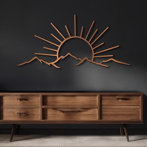 Pu&ograve; includere: Un'opera d'arte da parete in legno raffigurante un'alba su una catena montuosa. I raggi del sole si estendono verso l'esterno da dietro le montagne. L'opera d'arte &egrave; di colore marrone caldo ed &egrave; montata su una parete grigio scuro.