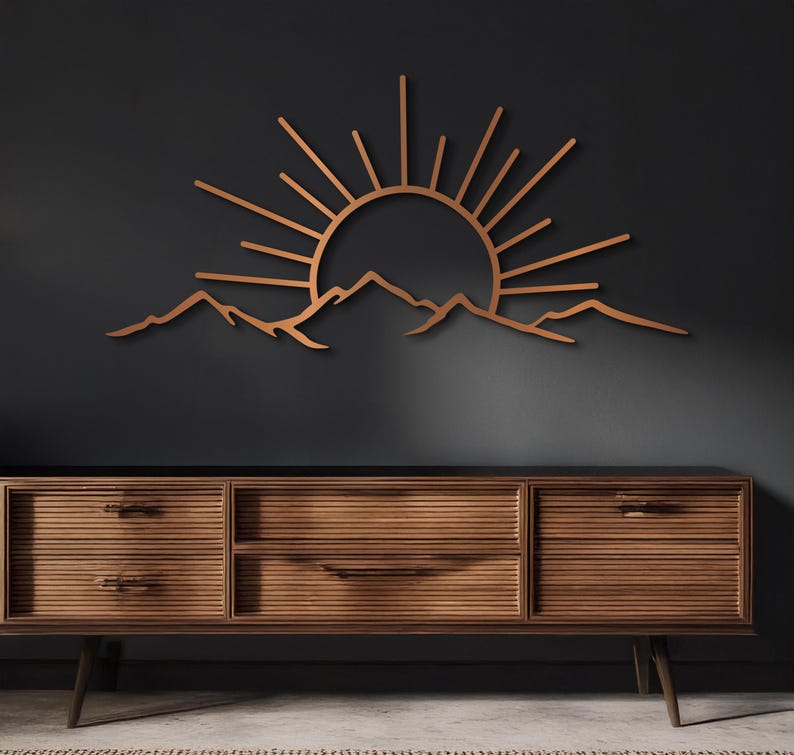 Pu&ograve; includere: Un'opera d'arte da parete in legno raffigurante un'alba su una catena montuosa. I raggi del sole si estendono verso l'esterno da dietro le montagne. L'opera d'arte &egrave; di colore marrone caldo ed &egrave; montata su una parete grigio scuro.