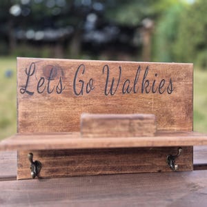 May include: A rectangular brown stained wooden wall organizer featuring a small shelf and two metal hooks. The text "Lets Go Walkies" is engraved in black cursive lettering on the upper portion of the wooden board above the shelf.