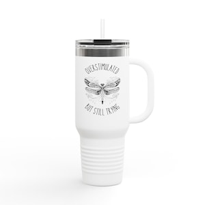 May include: White travel mug with a handle and a straw. The mug features the text "OVERSTIMULATED BUT STILL TRYING" around a detailed dragonfly illustration. The mug has a silver lid and a white handle.