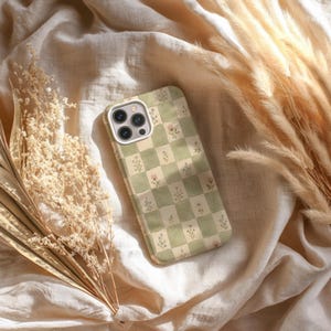May include: A phone case with a green and cream checkered pattern, adorned with small floral illustrations. The case has a white border around the camera and is resting on a cream-colored fabric, with dried plants in the background.