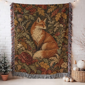May include: A rectangular woven tapestry throw blanket featuring a central fox surrounded by detailed botanical illustrations of oak leaves, red berries, thistles, and flowers in earthy tones, finished with a multi-colored fringed edge.
