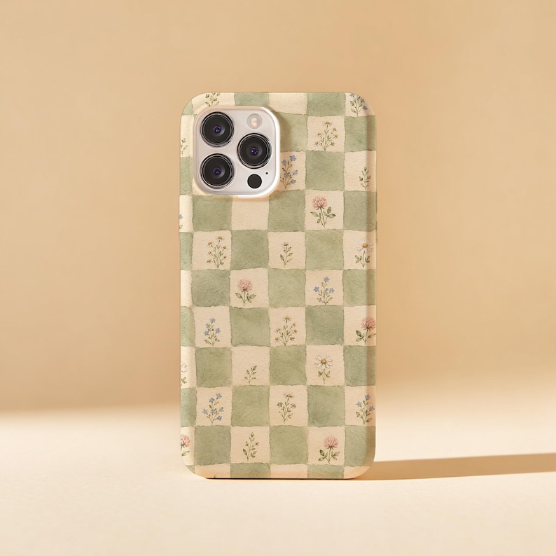 May include: A phone case with a green and white checkered pattern, adorned with small floral illustrations. The case has a camera cutout and a smooth finish, offering a blend of style and protection.