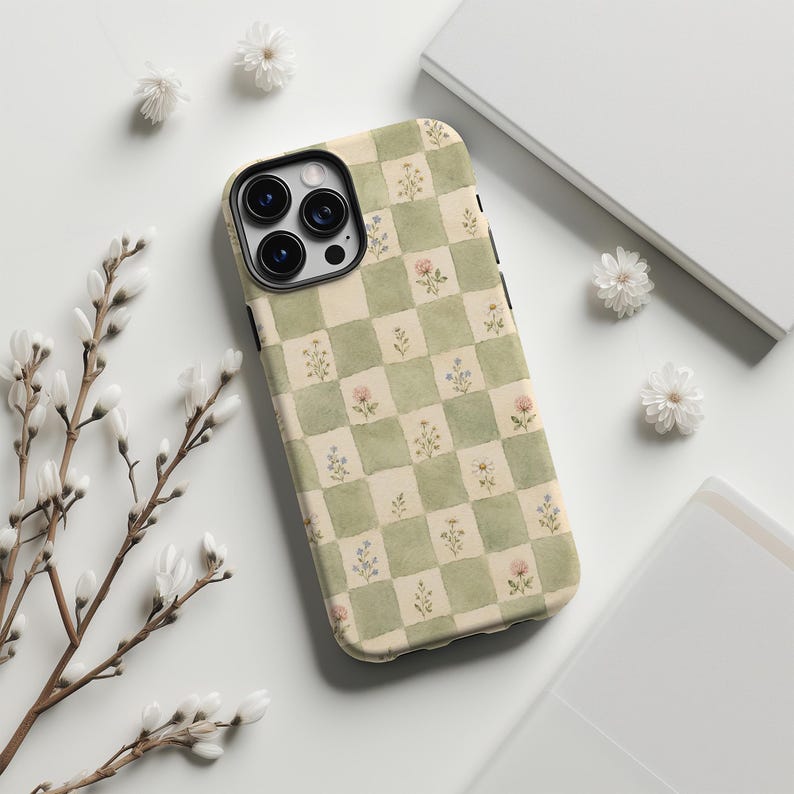 May include: A phone case with a green and cream checkered pattern, adorned with delicate floral illustrations. The case has a black camera module and a smooth, glossy finish. The design evokes a vintage, cottagecore aesthetic.
