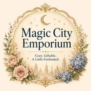 May include: A circular gold-toned frame surrounds the text Magic City Emporium, Cozy. Giftable. A Little Enchanted. The design features a crescent moon, stars, a city skyline silhouette, pink roses, and blue hydrangea flowers on a cream background.