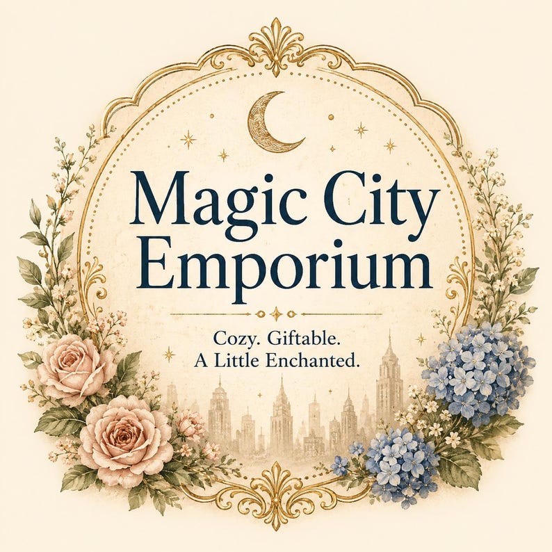 May include: A circular gold-toned frame surrounds the text Magic City Emporium, Cozy. Giftable. A Little Enchanted. The design features a crescent moon, stars, a city skyline silhouette, pink roses, and blue hydrangea flowers on a cream background.