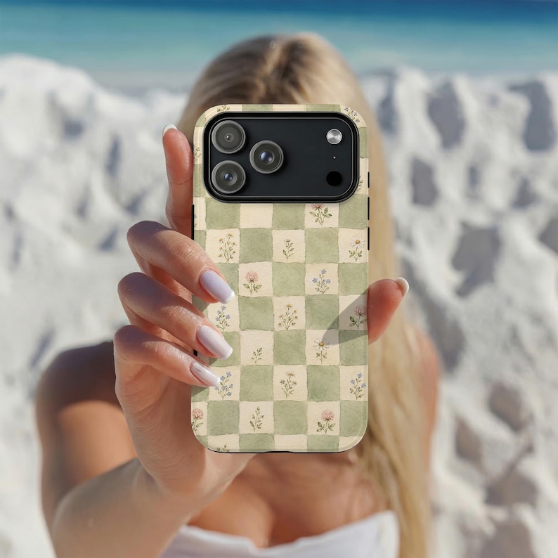 May include: A phone case with a green and cream checkered pattern, adorned with delicate floral illustrations. The case has a black camera module and is held up in front of a blurred background of a beach.
