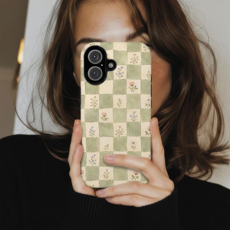 May include: A phone case with a checkered pattern of light green and cream squares. Each square features a small floral design in pink, blue, and white. The case has a camera lens cutout and is held in front of a person.