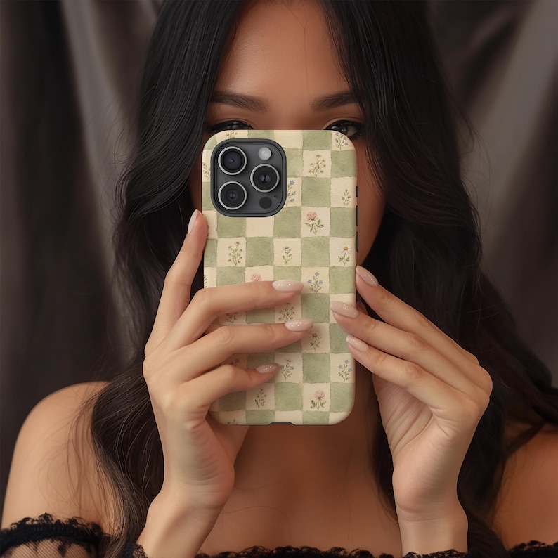 May include: A phone case with a green and cream checkered pattern, adorned with small floral illustrations. The case is held up in front of a person's face. The case has a camera module with three lenses.