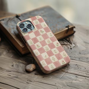 May include: A smartphone case featuring a chequered pattern of dusty pink and cream squares. Each square is decorated with a small, delicate floral illustration of tiny wildflowers and buds. The case has a cutout for a triple-lens camera system.