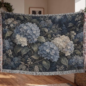 May include: A woven throw blanket featuring a dark navy blue background with a detailed floral pattern of light blue and white hydrangea clusters and green leaves. The rectangular blanket has a decorative fringed edge on all four sides.
