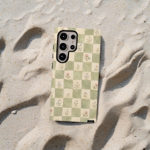 May include: A phone case with a green and white checkered pattern, adorned with small watercolor floral designs in pink and blue. The case has a camera cutout and is set against a sandy background.
