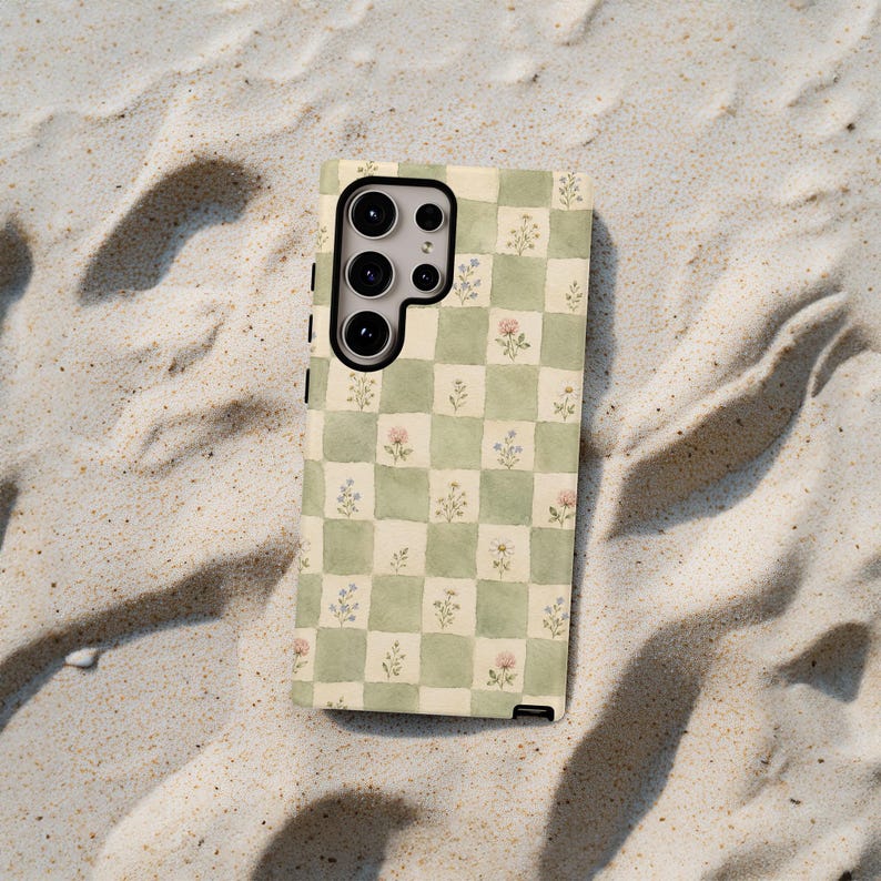 May include: A phone case with a green and white checkered pattern, adorned with small watercolor floral designs in pink and blue. The case has a camera cutout and is set against a sandy background.