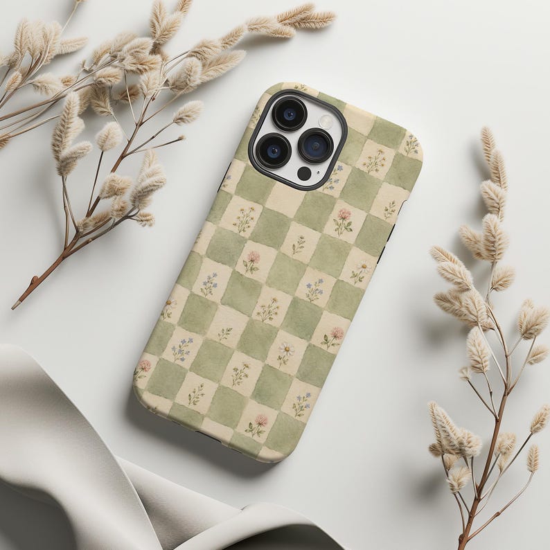 May include: A phone case with a green and cream checkered pattern, adorned with small floral illustrations. The case has a camera cutout and is surrounded by dried floral stems.
