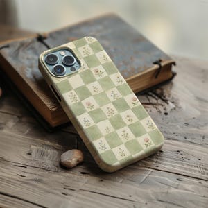 May include: A phone case with a green and white checkered pattern, adorned with small floral illustrations. The case is made of a durable material and is designed to fit a smartphone. The case is resting on a wooden surface.