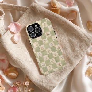 May include: A phone case with a green and white checkered pattern, adorned with small floral illustrations. The case is resting on a beige fabric surface, surrounded by scattered flower petals. The phone case has a black camera module.