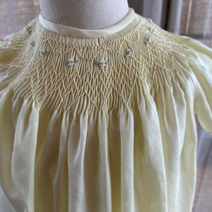 May include: A pale yellow smocked dress with short sleeves. The bodice features intricate smocking with delicate floral embroidery. The fabric is gathered below the smocked yoke, creating a flowing, comfortable silhouette. The dress is made of a soft, lightweight material.