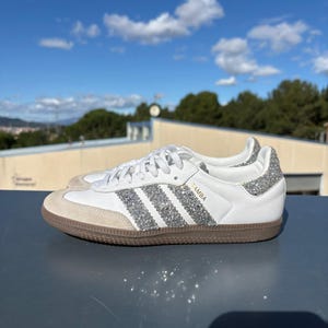 Könnte beinhalten: Weiße Samba-Sneaker aus Leder mit silbernen Glitzerakzenten an der Ferse und drei Streifen. Die Sneaker haben eine Zehenkappe aus beigem Wildleder und eine braune Gummisohle. Das Wort "SAMBA" ist goldfarben auf der Seite aufgedruckt.