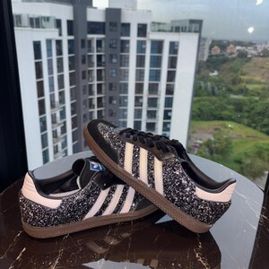 May include: Black glitter sneakers with white stripes and a brown sole. The shoes have a black tongue and laces, and a white heel tab. The design is a classic style.