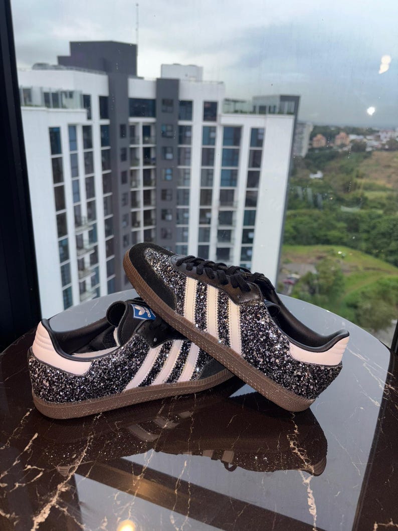 May include: Black glitter sneakers with white stripes and a brown sole. The shoes have a black tongue and laces, and a white heel tab. The design is a classic style.