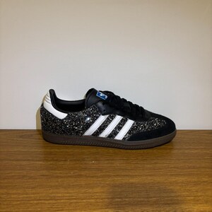 May include: Black and silver glitter Adidas Samba trainers with white stripes and a brown sole. The shoe has a black tongue and laces, and a white heel. The Adidas logo is visible on the tongue.