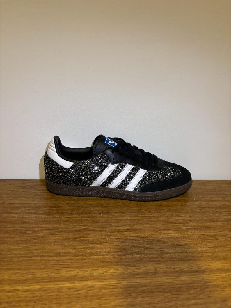 May include: Black and silver glitter Adidas Samba trainers with white stripes and a brown sole. The shoe has a black tongue and laces, and a white heel. The Adidas logo is visible on the tongue.