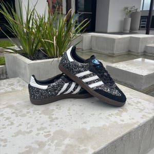 May include: Black glitter trainers with white stripes and a brown sole. The shoes feature a classic design with a blue logo on the tongue and white detailing on the heel. The trainers are a stylish footwear option.