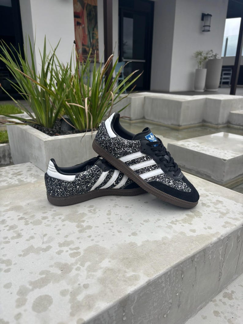 May include: Black glitter trainers with white stripes and a brown sole. The shoes feature a classic design with a blue logo on the tongue and white detailing on the heel. The trainers are a stylish footwear option.