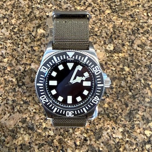 Peut inclure: Une montre-bracelet en acier inoxydable avec un cadran noir, des index g&eacute;om&eacute;triques blancs et des aiguilles blanches. La montre pr&eacute;sente une lunette rotative noire avec des marquages num&eacute;riques blancs et un bracelet en tissu vert olive.