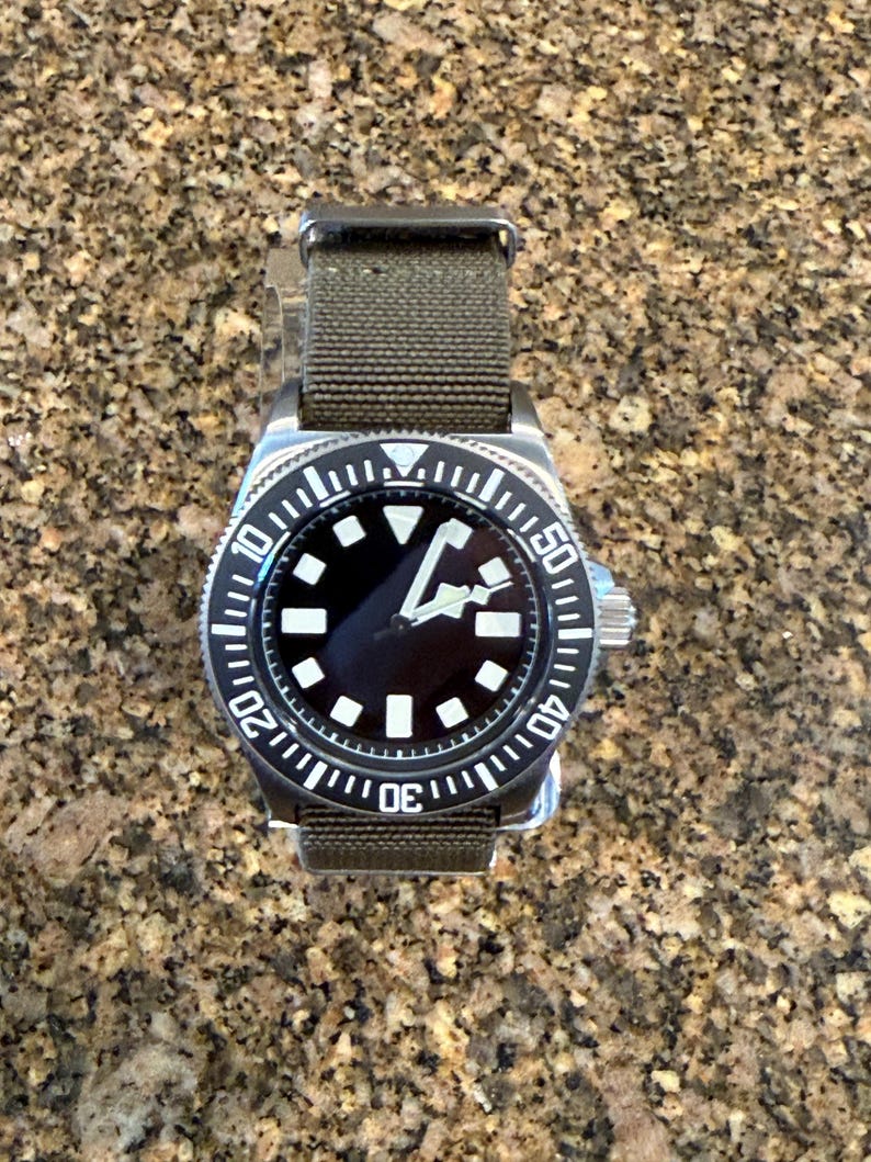 Peut inclure: Une montre-bracelet en acier inoxydable avec un cadran noir, des index g&eacute;om&eacute;triques blancs et des aiguilles blanches. La montre pr&eacute;sente une lunette rotative noire avec des marquages num&eacute;riques blancs et un bracelet en tissu vert olive.