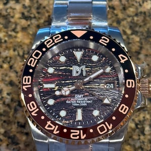 Puede incluir: Reloj de pulsera de acero inoxidable con bisel negro y burdeos. La esfera es oscura con detalles dorados y el texto "GMT AUTOMATIC WATER RESISTANT 100m:330ft". El reloj tiene una visualización de fecha a las 3 en punto.
