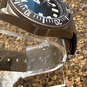 Peut inclure: Une montre-bracelet avec un bo&icirc;tier vert olive fonc&eacute; et un bracelet en tissu assorti. La montre pr&eacute;sente un cadran noir avec des index g&eacute;om&eacute;triques blancs, des aiguilles blanches et une lunette tournante noire avec des chiffres blancs.