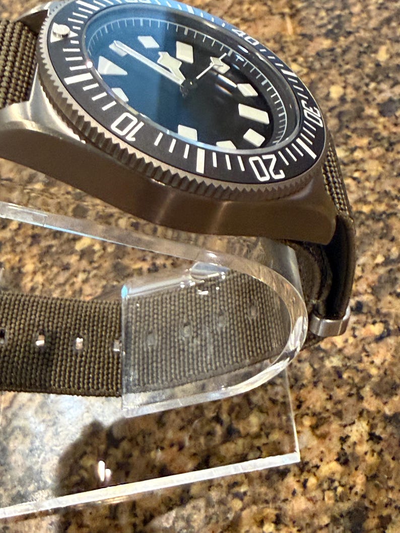 Peut inclure: Une montre-bracelet avec un bo&icirc;tier vert olive fonc&eacute; et un bracelet en tissu assorti. La montre pr&eacute;sente un cadran noir avec des index g&eacute;om&eacute;triques blancs, des aiguilles blanches et une lunette tournante noire avec des chiffres blancs.