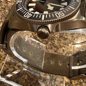 Peut inclure: Une montre-bracelet avec un bo&icirc;tier en m&eacute;tal bronze fonc&eacute; et une lunette rotative noire avec des marqueurs num&eacute;riques blancs. La montre pr&eacute;sente un cadran sombre avec des index lumineux g&eacute;om&eacute;triques et un bracelet en tissu vert olive.