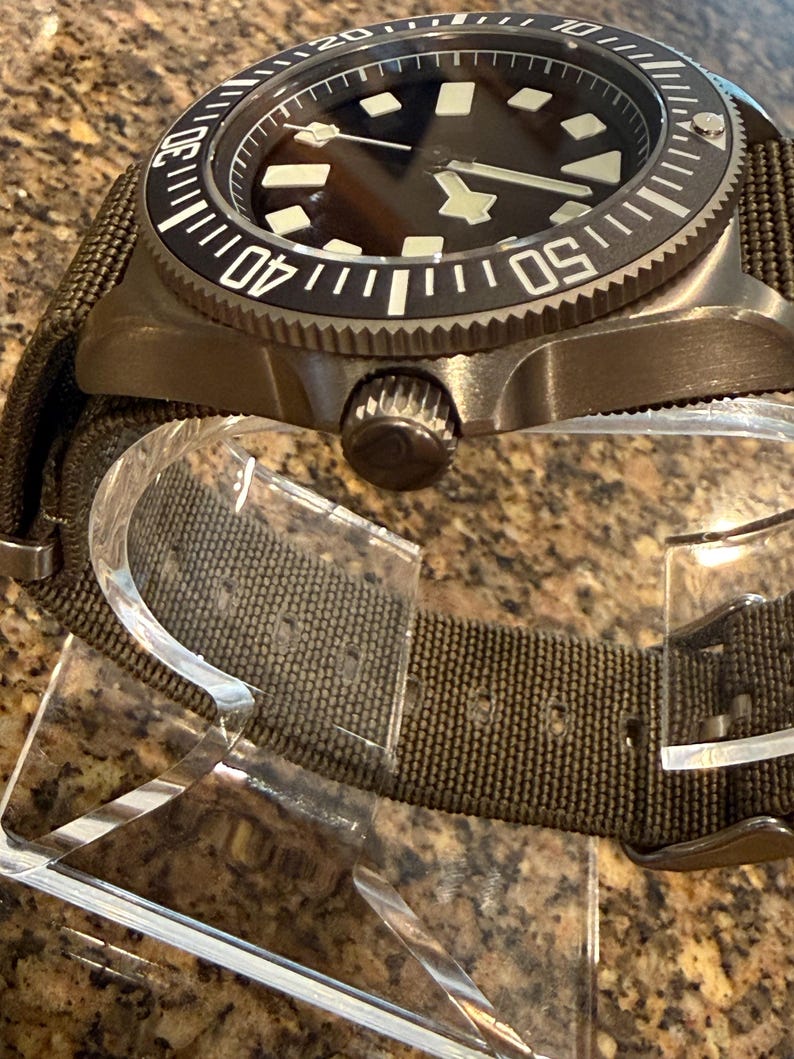 Peut inclure: Une montre-bracelet avec un bo&icirc;tier en m&eacute;tal bronze fonc&eacute; et une lunette rotative noire avec des marqueurs num&eacute;riques blancs. La montre pr&eacute;sente un cadran sombre avec des index lumineux g&eacute;om&eacute;triques et un bracelet en tissu vert olive.
