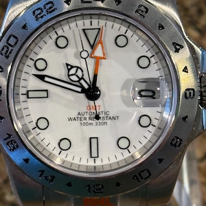 May include: A stainless steel wristwatch with a white face, black numerals, and a date window. The watch features a GMT hand with an orange arrow and is water-resistant up to 330 feet. The watch has a silver band and the text "GMT AUTOMATIC WATER REISTANT 100m:330ft".