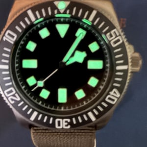 Peut inclure: Une montre-bracelet avec un cadran noir, des index lumineux verts et des aiguilles assorties. La montre pr&eacute;sente une lunette rotative noire avec des chiffres blancs et un bracelet en tissu marron. Le design inclut un marqueur triangulaire &agrave; douze heures et une trotteuse avec une petite pointe lumineuse carr&eacute;e.
